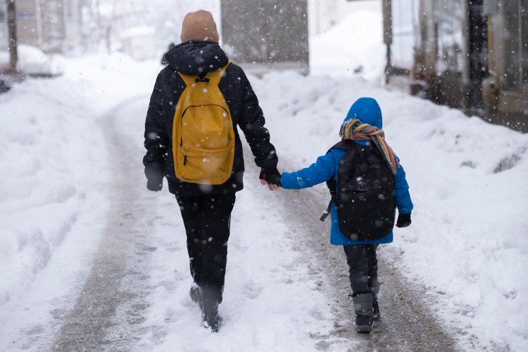 Et større barn med skolesekk leier et mindre barn med skolesekk på snødekt vei.